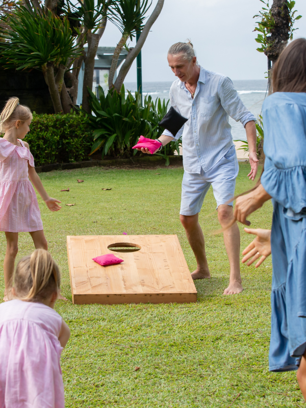 A family playing a game of Cornhole on the lawn in a tropical setting. One team is a little girl and her father, and the other is another little girl and her mother. The mother is throwing a bean bag into the cornhole board. 